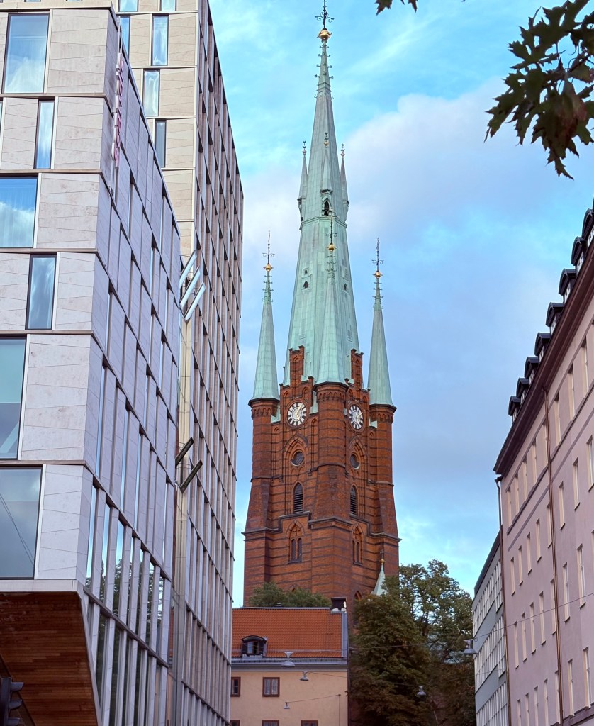 View of a tall church steeple with green roofs and red brick walls, framed by modern buildings on either side.