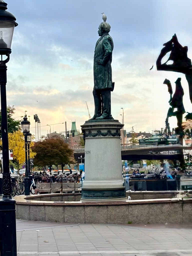 A statue of a man in a coat standing on a pedestal, with a seagull perched on his head, in a public square surrounded by trees and buildings.